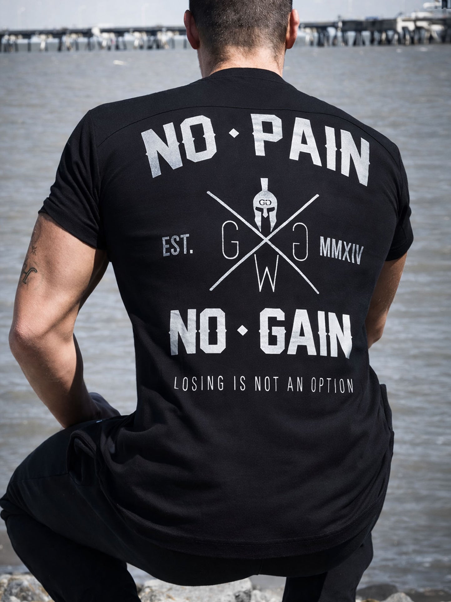 Schwarzes Gym Generation T-Shirt mit grossem „No Pain No Gain“-Rückenprint, getragen von hinten am Strand.