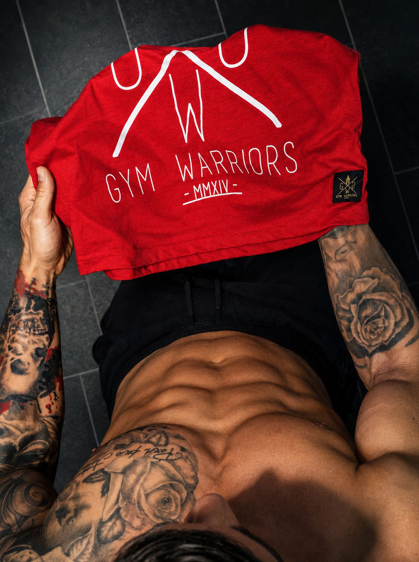 Gym Generation Stringer Gym Warriors in Rot – Detailansicht des weissen Gym-Warriors-Schriftzugs mit gewebtem Marken-Label am Saum, ärmelloses Muskelshirt für Herren