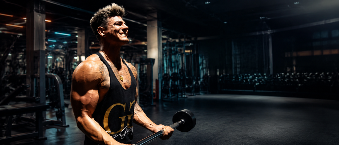 Athlet trägt einen schwarzen Gym Warriors Stringer beim Bizeps Training mit Langhantel in einem dunklen Premium-Gym.