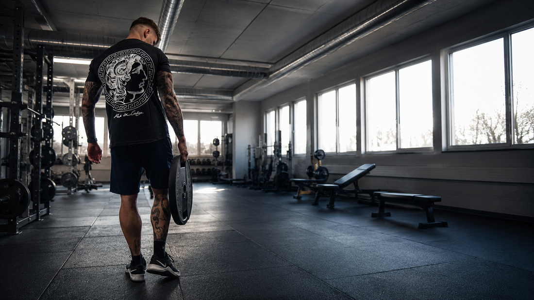 Athlet im Gym trägt schwarzes Gym Generation Alexander T-Shirt mit grossem Rückenprint und hält eine Hantelscheibe beim Krafttraining.