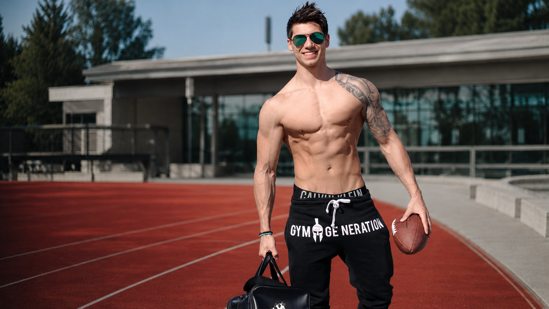 Athletischer Mann mit Sixpack trägt schwarze Gym Generation Trainingshose auf der Laufbahn mit Football und Sporttasche