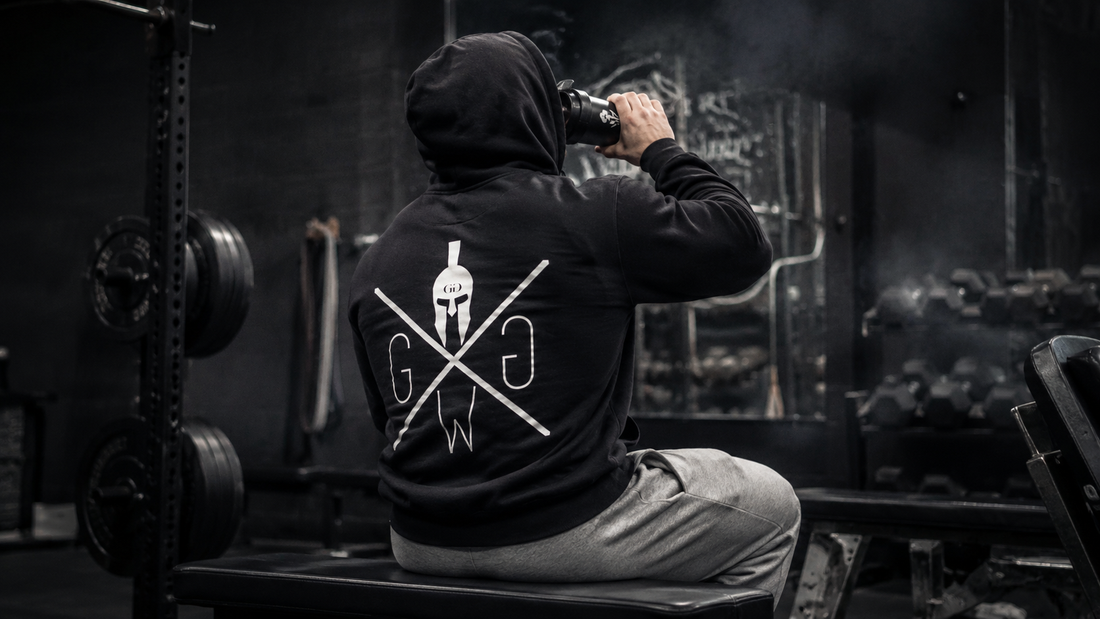 Mann trägt schwarzen Gym Warriors Hoodie im Gym und trinkt aus einem Shaker nach dem Training