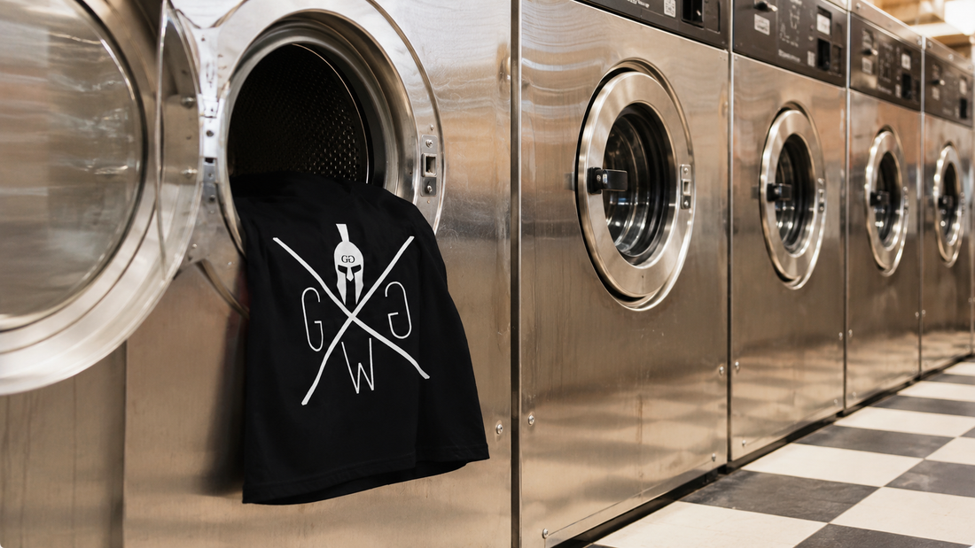 Schwarzes Gym Warriors Shirt hängt aus einer Waschmaschine im Waschsalon als Symbol für Pflege von Fitnessbekleidung