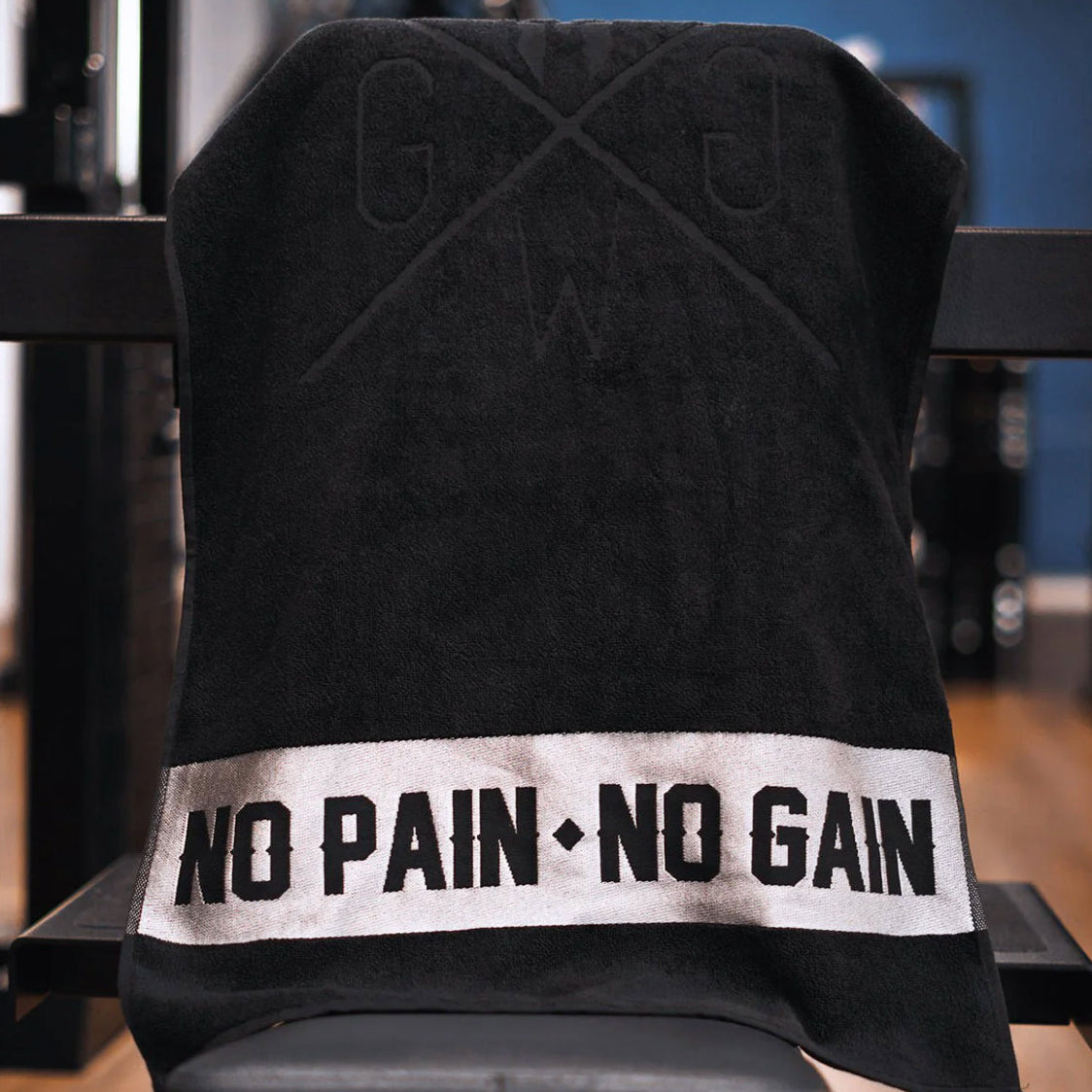 Schwarzes Gym Generation Sporthandtuch aus Baumwoll-Frottee mit gesticktem No Pain No Gain Schriftzug und eingewebtem GG-Logo, über einer Trainingsbank im Fitnessstudio, Format 50 × 100 cm