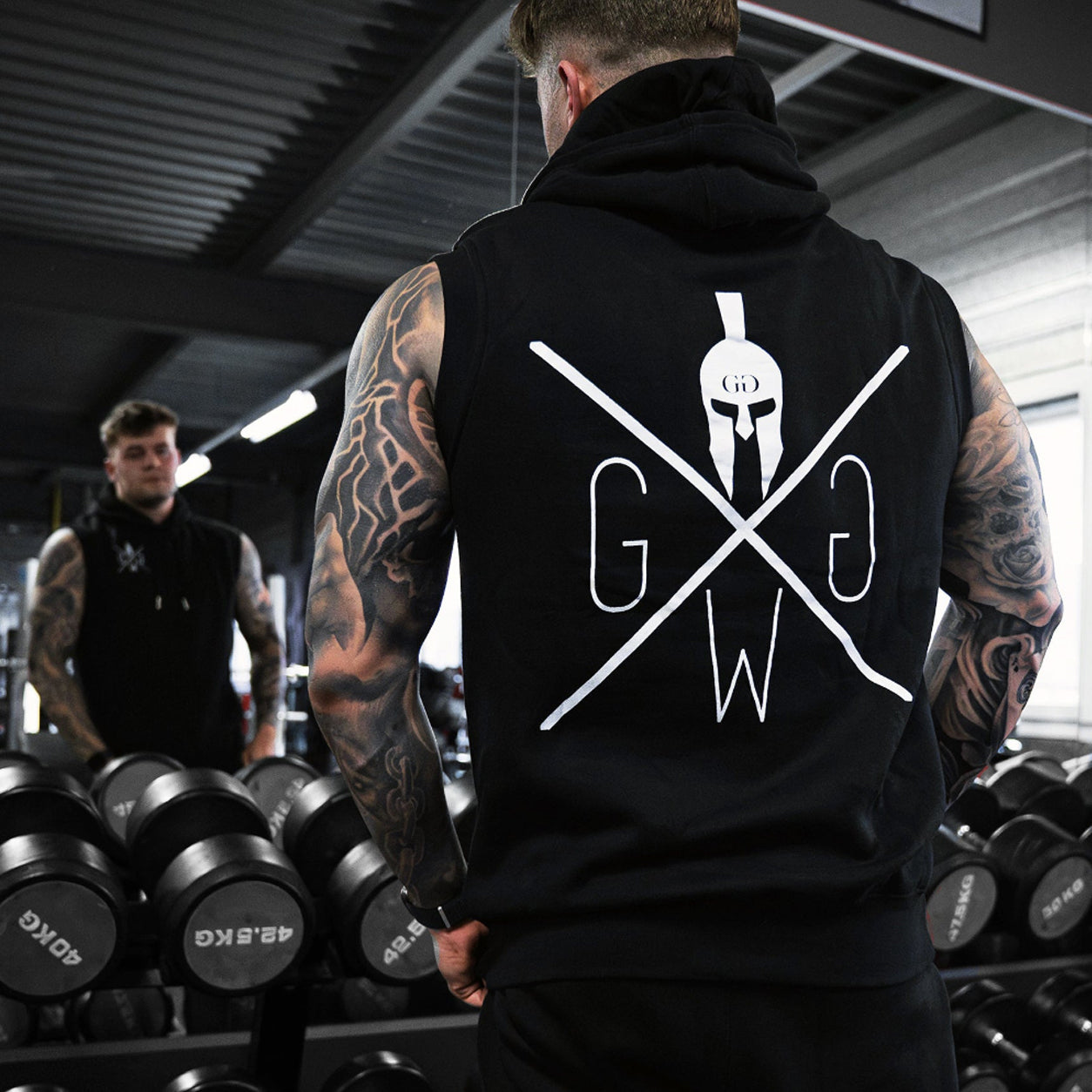 Bodybuilder trägt schwarzes Pump Cover mit Gym Warriors Logo am Hantelständer im Gym – Gym Generation