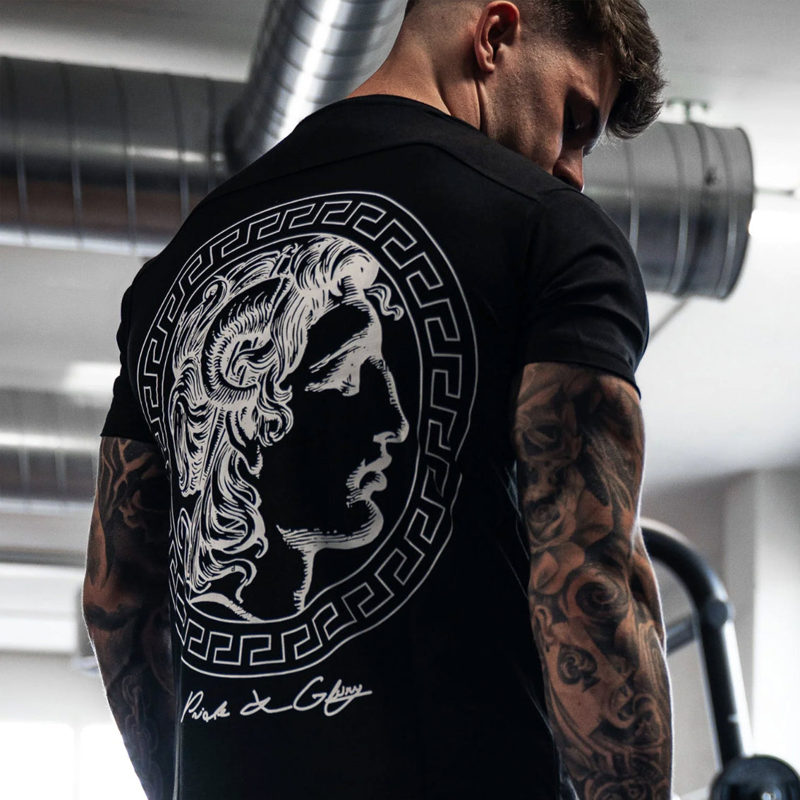 Mann mit Tattoos trägt schwarzes Gym Generation T-Shirt mit Alexander-the-Great-Backprint im griechischen Mäander-Kreis im Fitnessstudio — Athletic Fit, 100% Baumwolle, Pride & Glory Schriftzug