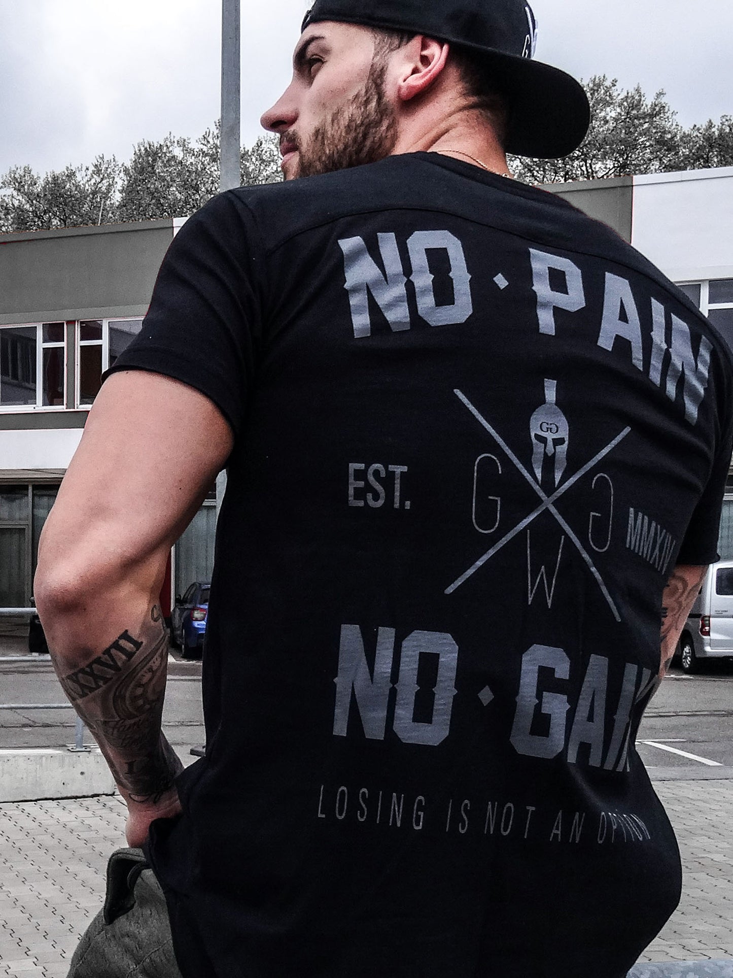 Rückenansicht des Gym T-Shirts 'No Pain No Gain' in Schwarz von Gym Generation, getragen im Freien, zeigt den markanten Druck mit dem Slogan 'No Pain No Gain' und 'Losing is Not an Option'.