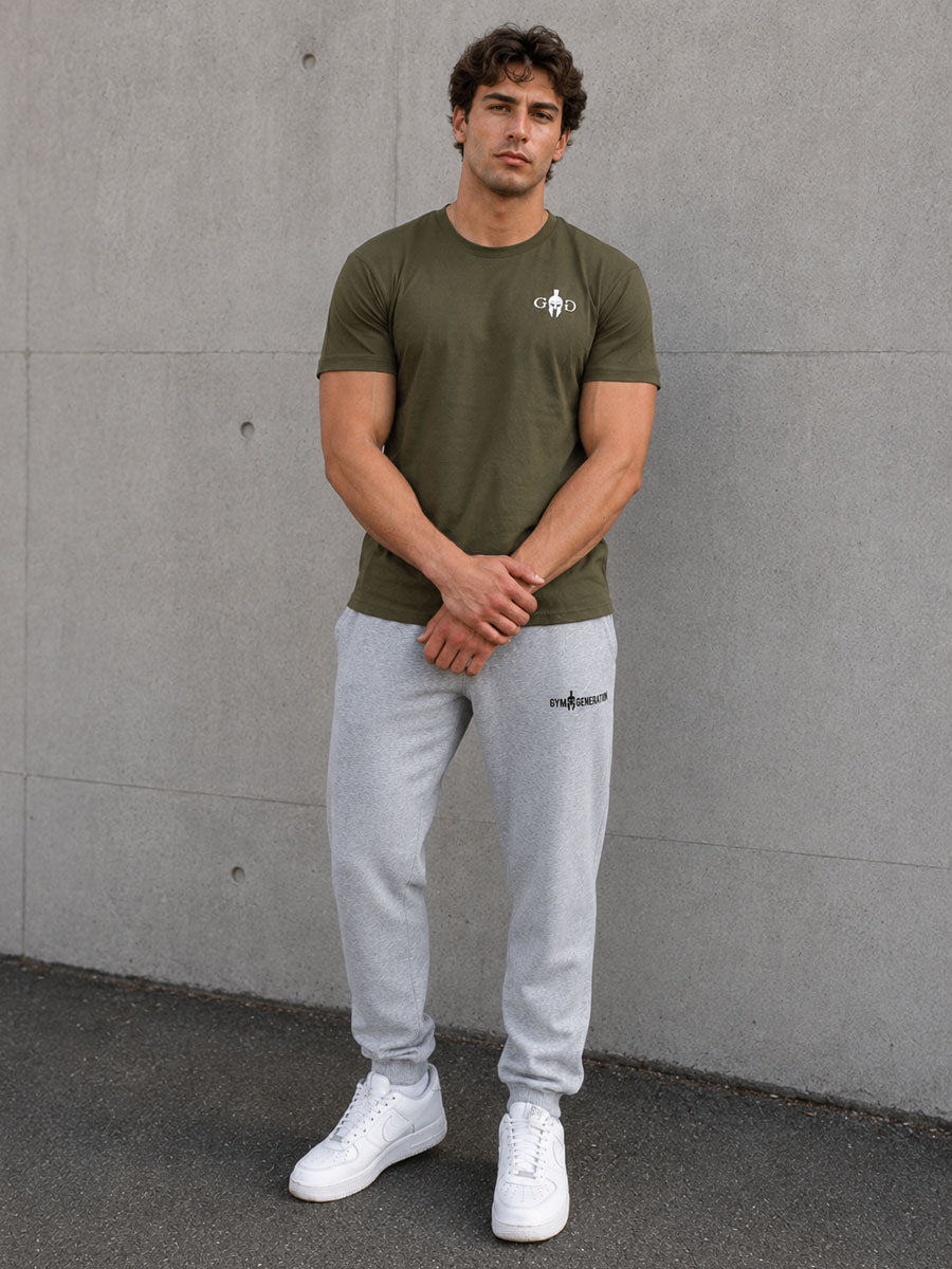Olives Fitness Shirt Herren von Gym Generation in Frontansicht, sportlicher Athletic Fit mit weissem Brustlogo, grauer Jogginghose und weissen Sneakers vor Betonwand
