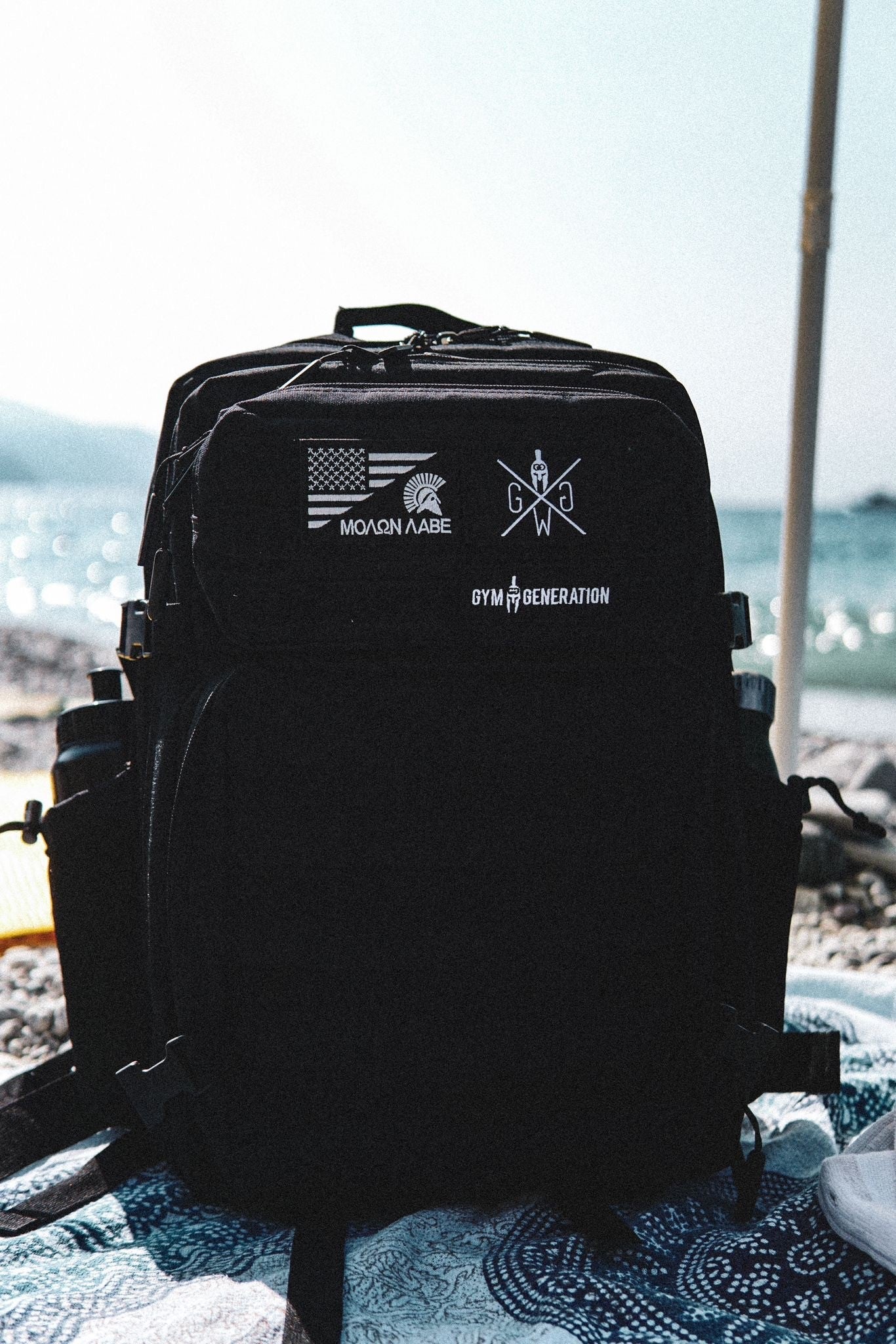 Schwarzer Gym Generation Rucksack mit verschiedenen Patches am Strand – stilvoll und praktisch, ideal für Fitness, Outdoor-Abenteuer und Reisen.