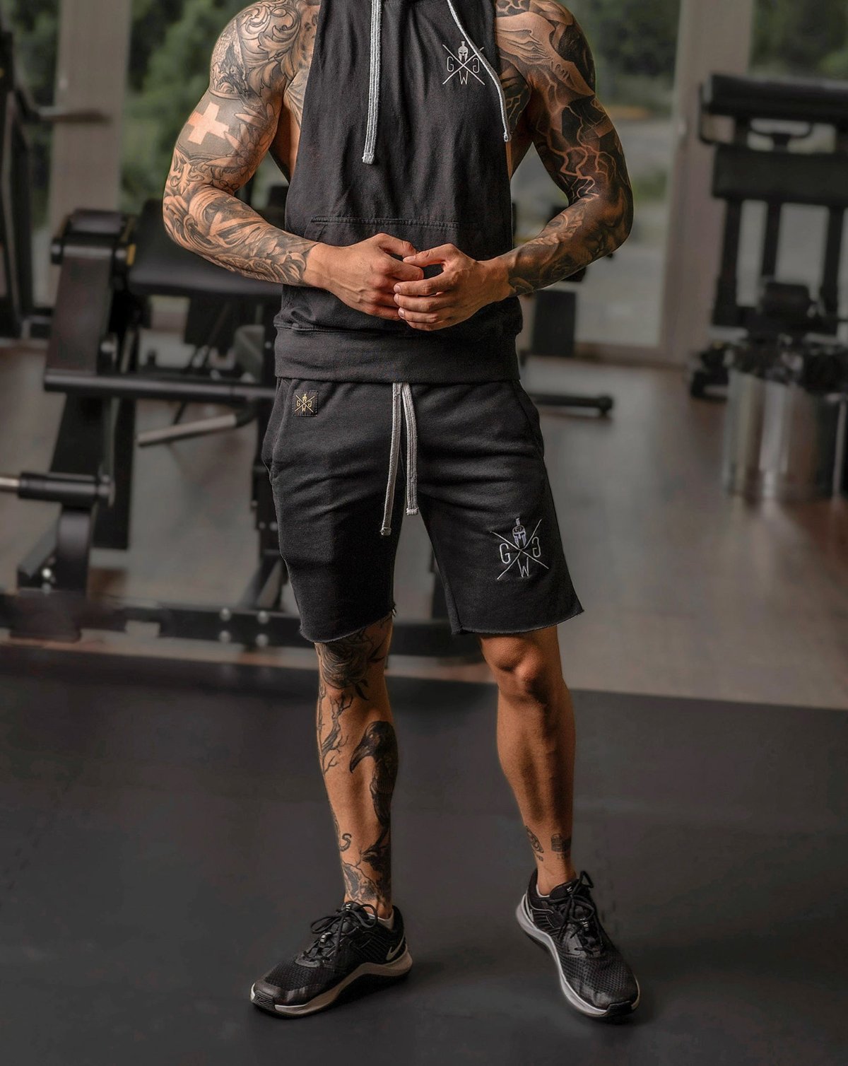 Tätowierter Mann in einem schwarzen Gym Generation Tanktop und Shorts mit Logo, kombiniert mit Sneakers, im Fitnessstudio während eines Trainings. Ideal für Bodybuilding, Krafttraining und sportliche Aktivitäten.