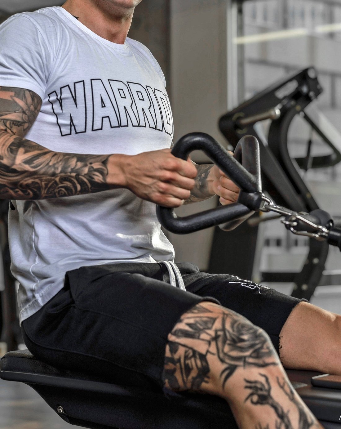 Tätowierter Mann im weißen "WARRIORS"-T-Shirt und schwarzen Gym Generation Shorts beim Training an einem Kabelzuggerät im Fitnessstudio. Ideal für Krafttraining, Bodybuilding und sportliche Aktivitäten.