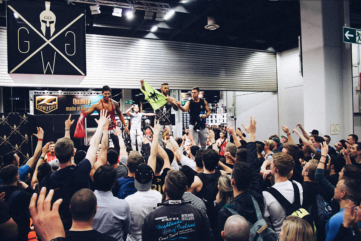 Gym Generation Athleten auf einer Fitnessmesse verteilen Sportbekleidung an begeisterte Fans, die jubelnd die Hände heben, symbolisiert die dynamische Markeninteraktion, Motivation und den starken Gemeinschaftssinn der Fitness-Community.