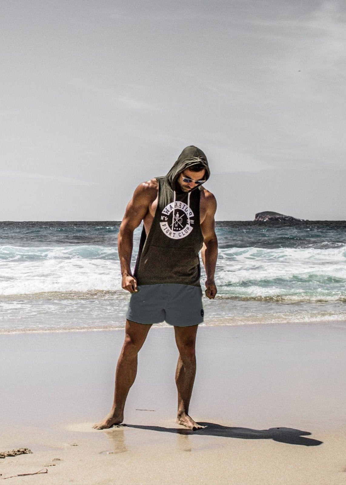 Muskulöser Mann in einem Gym Generation Hoodie-Tanktop in olive mit Logo und grauen Shorts, barfuß am Strand stehend, ideal für Fitness, Freizeit und sportlichen Lifestyle.