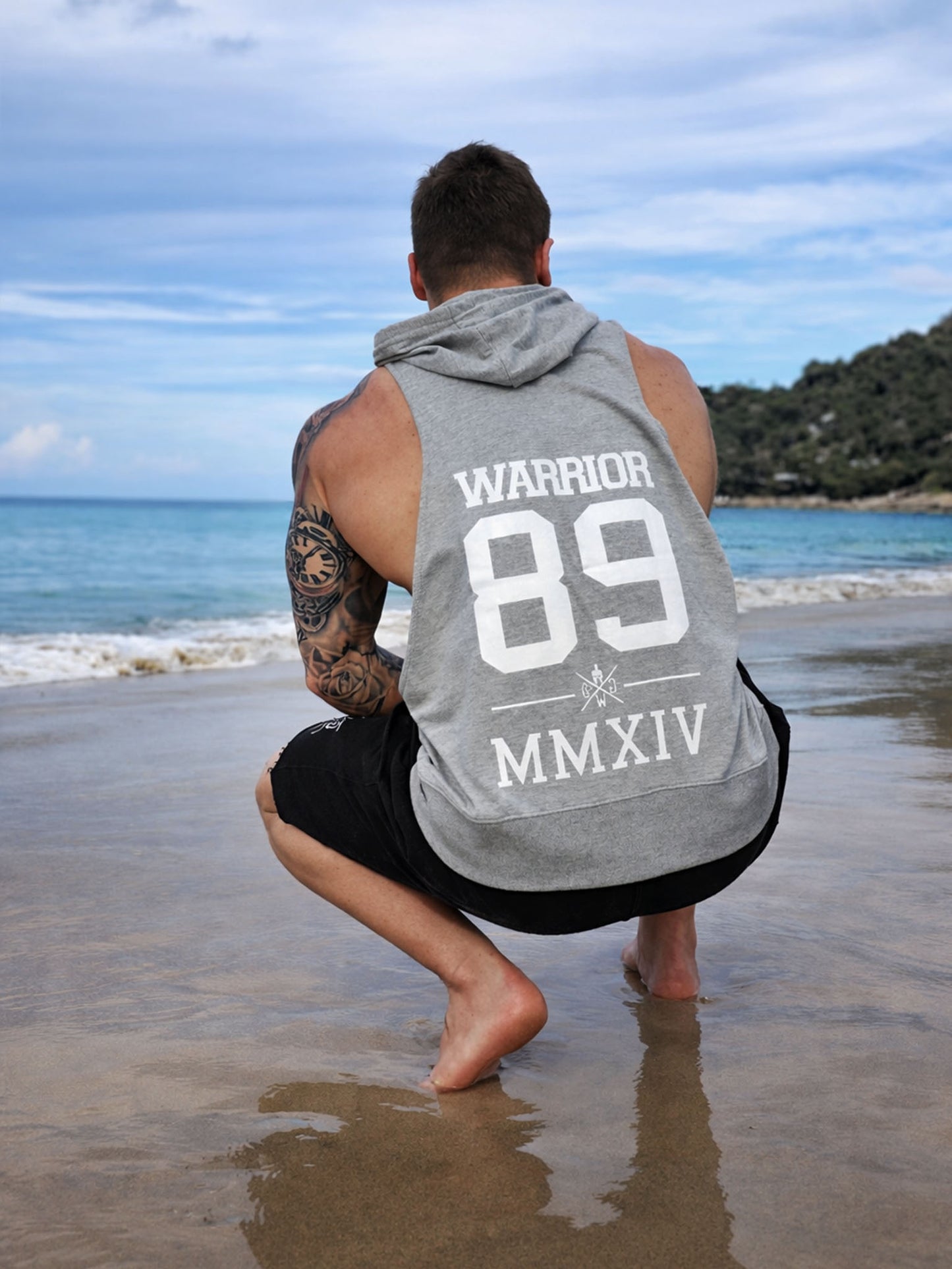 Graues Kapuzen Tank Top für Herren von Gym Generation in Outdoor-Rückansicht am Strand – ärmelloser Fitness Hoodie mit großem Warrior 89 Rückenprint
