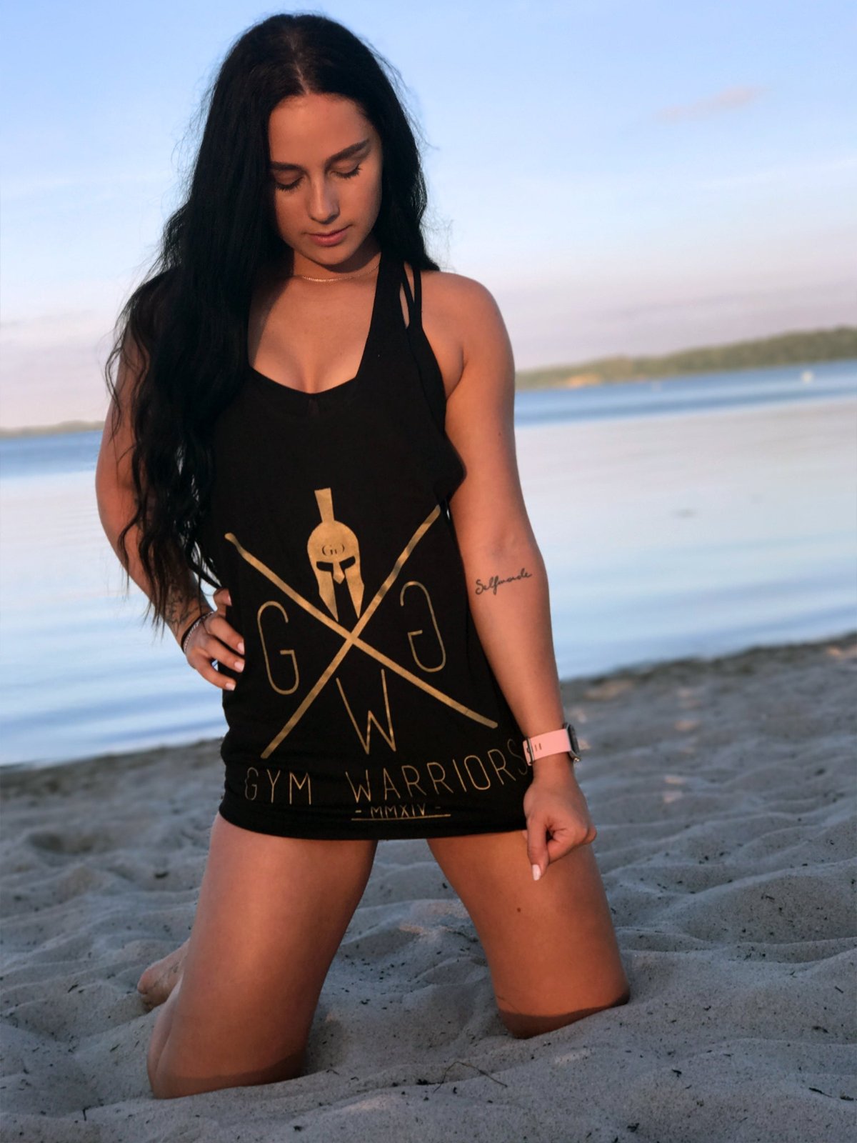 Schwarzes Stringer Tank Top mit goldenem Gym Warriors Print, getragen von weiblichen Model am Strand – perfekt für Fitness, Freizeit und sommerlichen Style.