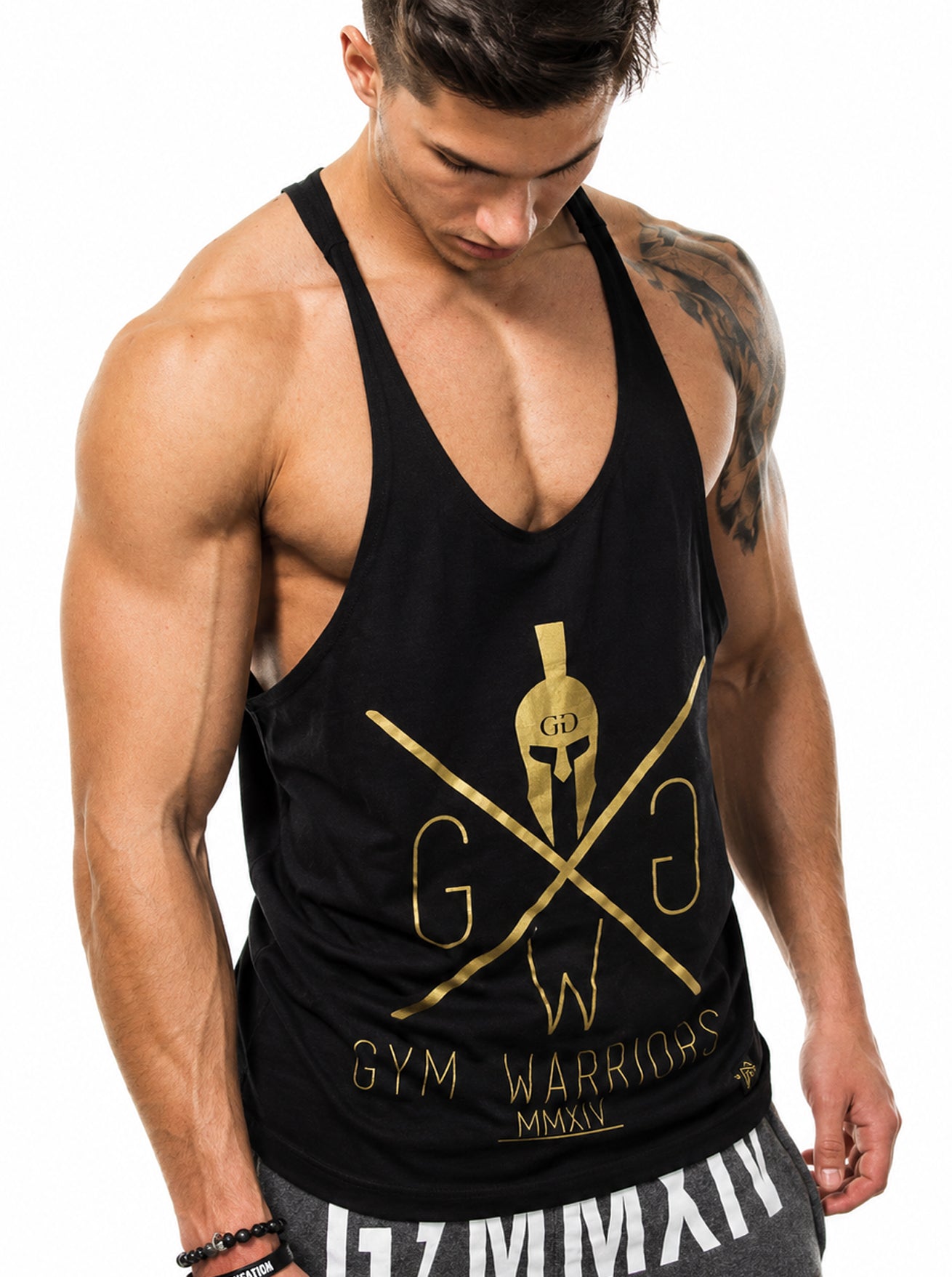 Gym Generation Stringer Gym Warriors in Schwarz mit goldenem Spartaner-Print am Model – ärmelloses Trainingsshirt für Herren, kombiniert mit grauen Joggern im Racerback-Schnitt