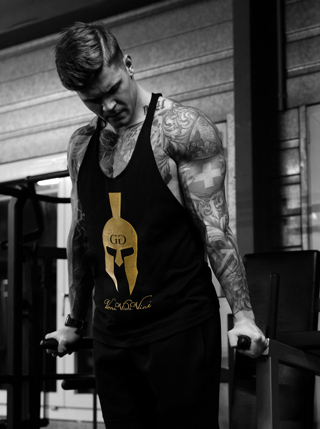 Tätowierter Athlet trainiert Dips am Barren im schwarzen Gym Generation Stringer Veni Vidi Vici – goldener Spartaner-Helm mit GG-Logo und Veni-Vidi-Vici-Schriftzug, ärmelloses Bodybuilding-Shirt für Herren mit tiefem Racerback-Rücken
