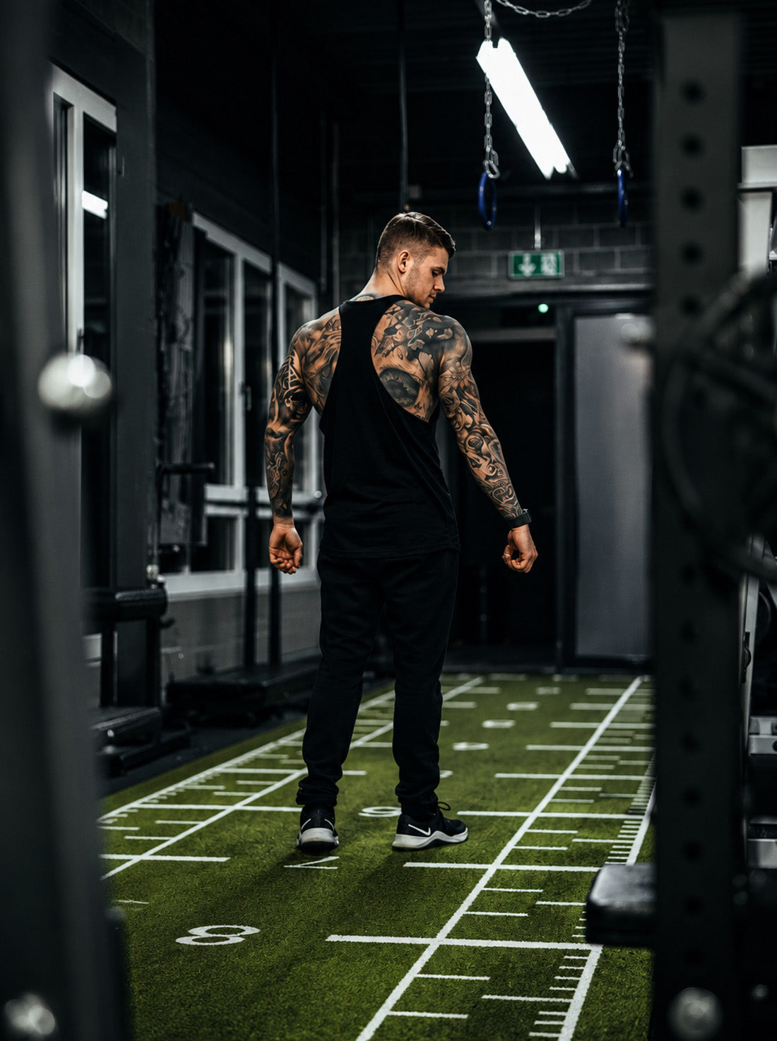 Gym Generation Stringer Veni Vidi Vici in Schwarz von hinten im Gym – Athlet auf der Sled-Track, tiefer Racerback-Schnitt, ärmelloses Trainingsshirt für Herren