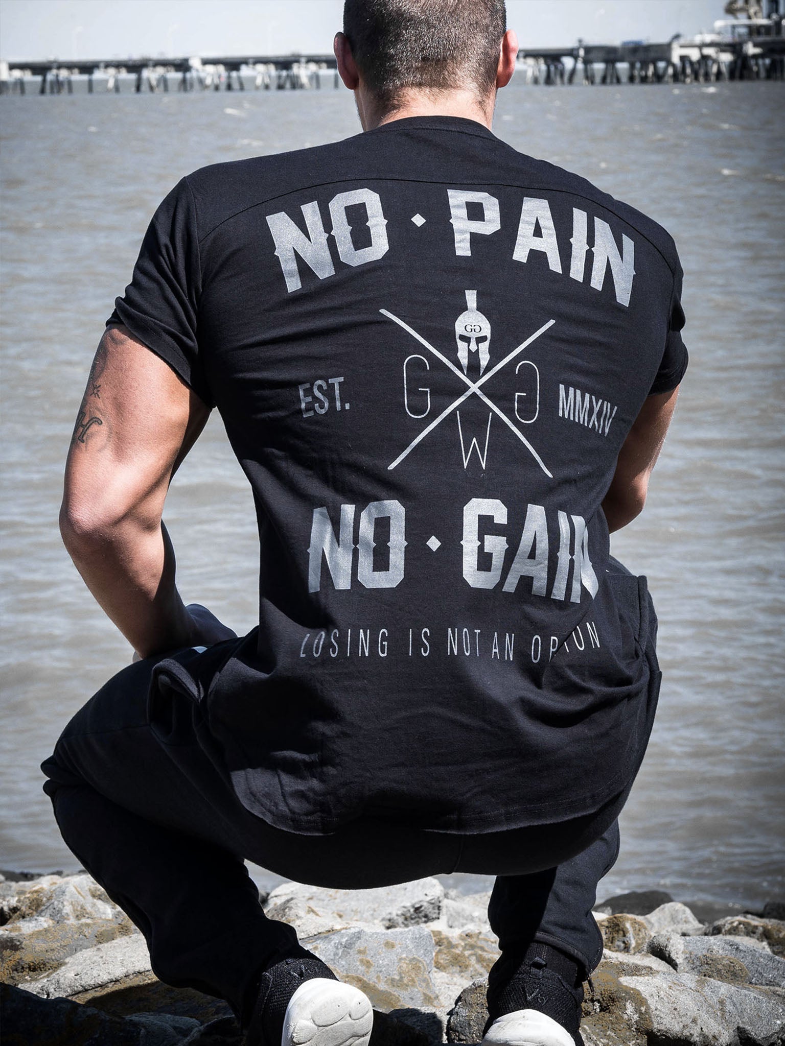 Gym T-Shirt 'No Pain No Gain' in Schwarz von Gym Generation, getragen an einem Strand mit dem markanten Druck 'No Pain No Gain' und 'Losing is Not an Option'.