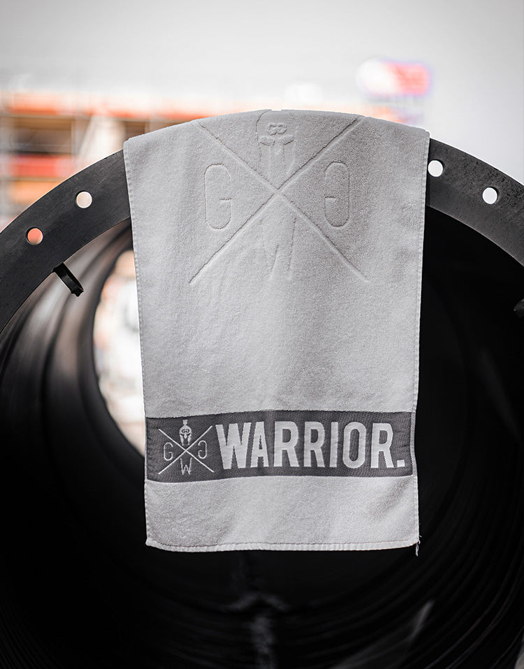 Off-white Fitness-Handtuch von Gym Generation mit geprägtem Kreuz-Logo und „WARRIOR“-Schriftzug, hängend an schwarzer Hantelstange im Gym.