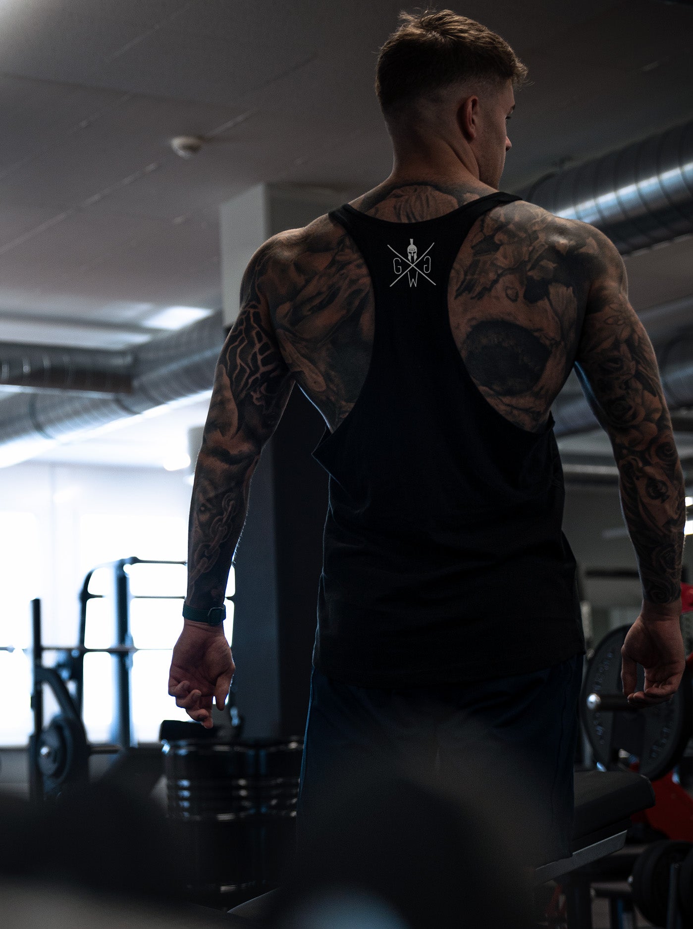 Rückansicht eines tätowierten muskulösen Mannes in einem schwarzen Tanktop mit Gym Generation Logo beim Training in einem Fitnessstudio mit moderner Ausstattung.