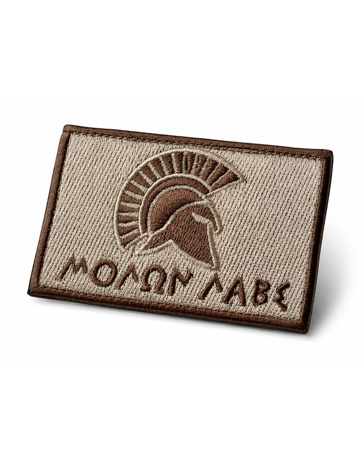 Molon Labe Patch Bronze schräg fotografiert – dichte Stickerei, umnähte braune Kante und Klett-Rückseite, ton-in-ton in Sand- und Bronzetönen.
