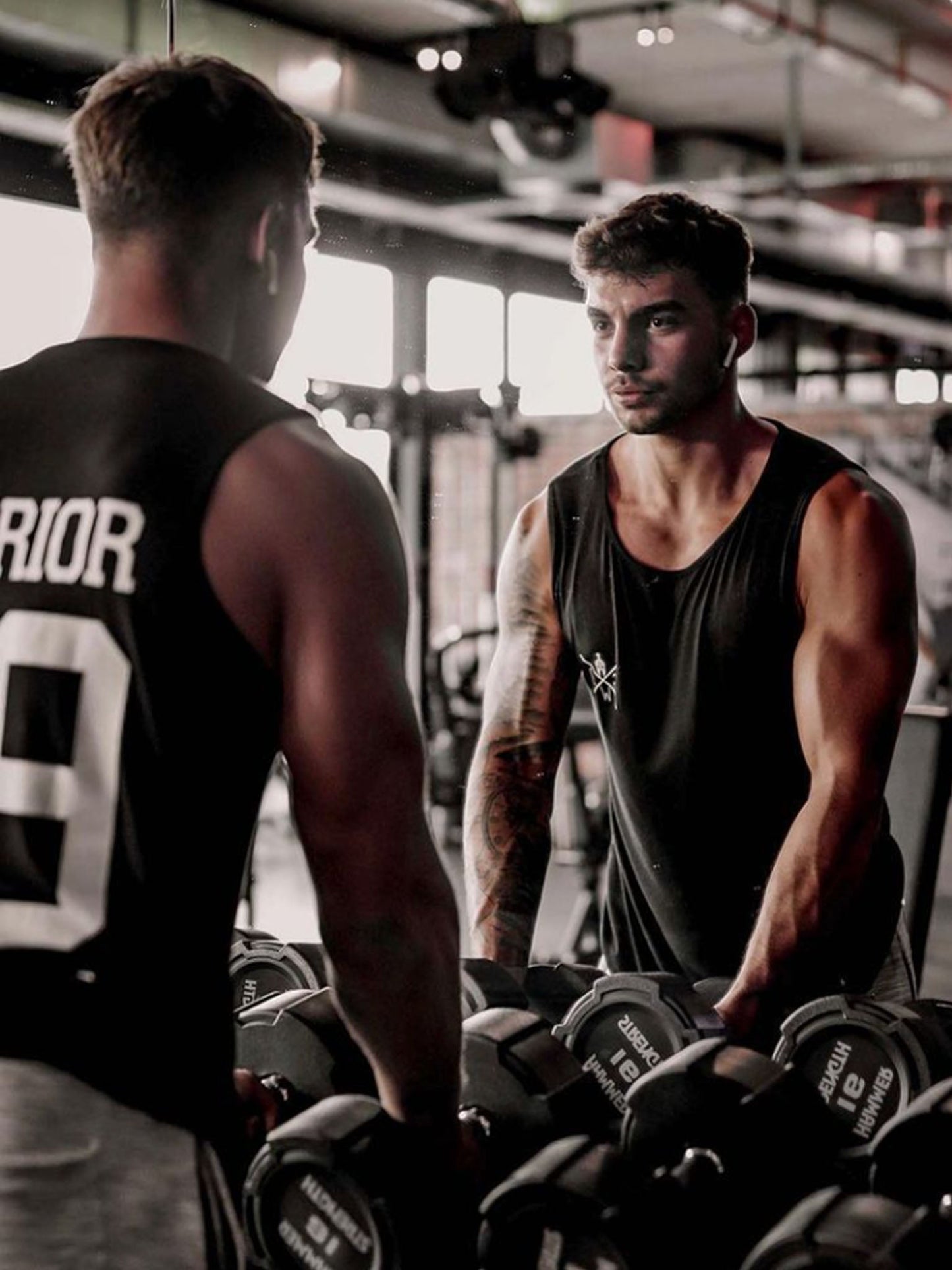 Athlet in einem Fitnessstudio, trägt das Gym Generation Muscle Tank Top in Schwarz, betrachtet sich konzentriert im Spiegel zwischen Hanteln.