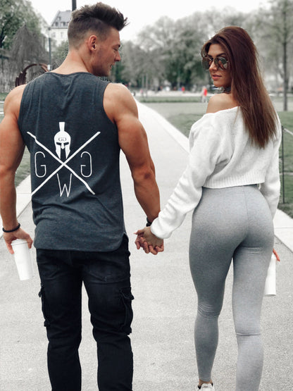 Fitness-Paar im Park: Der Mann trägt das Gym Generation Fitness Tank Top Warrior in Dunkelgrau mit Spartaner-Logo. Beide halten Becher und gehen Hand in Hand.